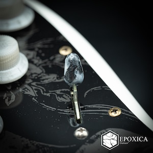 May include: A black guitar pickguard with a white floral pattern, featuring a marbled grey and white pickup selector switch tip, white control knobs, and gold-colored screws. The logo EPOXICA Guitars is printed in white in the bottom corner.
