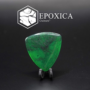 May include: A green guitar plectrum with black marbling, displayed on a small, dark metal stand. The plectrum has a triangular shape with rounded edges. The brand name "EPOXICA Guitars" is displayed in white text above the pick.