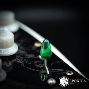 May include: A vibrant green, teardrop-shaped guitar pickup selector switch tip with a glossy finish, mounted on a black guitar body featuring a subtle etched pattern. The logo EPOXICA Guitars is visible in the bottom right corner of the image.