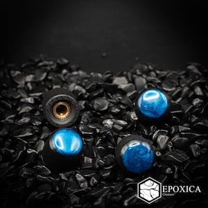 May include: Three cylindrical guitar knobs with matte black sides and glossy, swirling blue resin tops. One knob is inverted to show the brass-coloured, serrated metal insert. The text EPOXICA Guitars is printed in white in the bottom right corner.