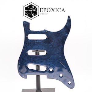 May include: A blue and black marbled guitar pickguard with a glossy finish, featuring three pickup cutouts and several screw holes. Above the pickguard, the logo for EPOXICA Guitars is displayed, consisting of a hexagon icon and text.