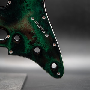 May include: A guitar pickguard with a swirling green and black pattern. The pickguard has several holes for screws and pickups, and a slot for the tremolo bar. The edges are smooth and rounded.