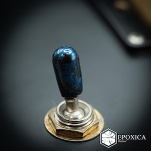 May include: A dark blue metallic guitar toggle switch tip with a glossy finish, mounted on a silver-colored metal base with a hexagonal nut. The component is attached to a matte black surface, featuring the logo text EPOXICA Guitars in the corner.