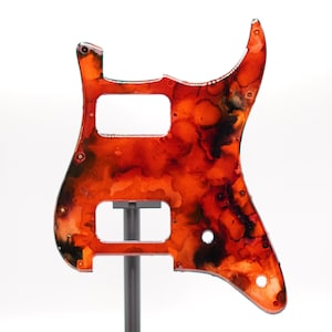 Puede incluir: Un golpeador de guitarra con un dise&ntilde;o abstracto vibrante en tonos rojos, naranjas y negros. El golpeador tiene recortes para pastillas y controles, y est&aacute; montado en un soporte. El dise&ntilde;o se asemeja a una acuarela.
