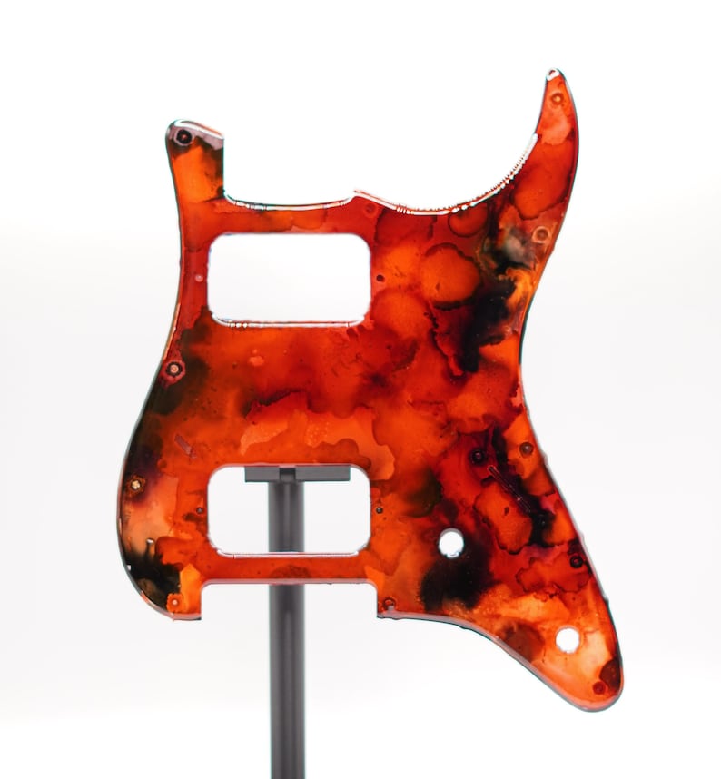 Puede incluir: Un golpeador de guitarra con un dise&ntilde;o abstracto vibrante en tonos rojos, naranjas y negros. El golpeador tiene recortes para pastillas y controles, y est&aacute; montado en un soporte. El dise&ntilde;o se asemeja a una acuarela.