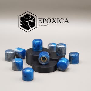 May include: A selection of blue and grey guitar knobs with a marbled design. Some knobs are placed on black circular platforms. The brand name "EPOXICA Guitars" is shown in black text.
