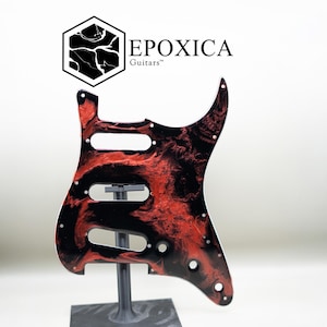 May include: A guitar pickguard featuring a swirling black and red marbled pattern. The piece includes three rectangular cutouts for pickups, a circular hole for a volume or tone knob, and several small screw holes. The logo EPOXICA Guitars is visible.