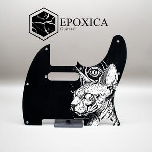 May include: Black guitar pickguard featuring a white Sphynx cat illustration and an eye. The pickguard has the text "EPOXICA Guitars" and a geometric logo in the upper left corner.