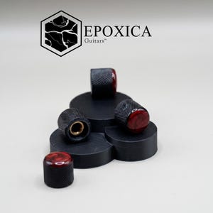 May include: A collection of black guitar knobs with a red and brown marbled top, arranged on a black platform. The knobs have a textured, ridged design. The logo "EPOXICA Guitars" is visible in the background.