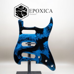 Peut inclure: Un corps de guitare avec un motif marbré bleu et noir. Le corps comporte des découpes pour les micros et les commandes. La marque "EPOXICA Guitars" est affichée dans le coin supérieur gauche.