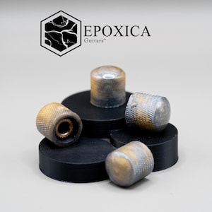 Op de afbeelding: Vier gitaarknoppen met een metallic afwerking, in tinten goud en zilver, worden weergegeven op zwarte cirkelvormige platforms. De knoppen hebben een textuur en een cilindrische vorm. De merknaam "EPOXICA Guitars" is zichtbaar.