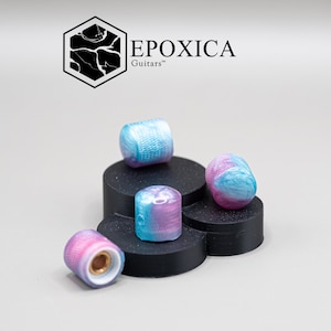 Op de afbeelding: Vier kleurrijke gitaarsnaren met een blauw, roze en paars gemarmerd ontwerp worden gepresenteerd op een zwart platform. De merknaam "EPOXICA Guitars" is zichtbaar in het bovenste deel van de afbeelding.