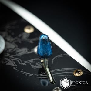 May include: A metallic blue guitar pickup selector switch tip with a glossy finish, mounted on a black guitar body featuring a subtle grey line pattern, gold-colored screws, and the white text logo EPOXICA Guitars in the bottom corner.