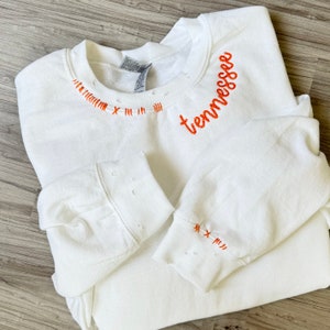 Może przedstawiać: Biała bluza z detalami w stylu vintage i pomarańczowym haftem. Słowo "tennessee" jest wyhaftowane zakrzywioną czcionką na g&oacute;rnej prawej piersi. Pomarańczowe szwy zdobią dekolt i mankiety.