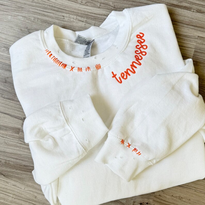 Może przedstawiać: Biała bluza z detalami w stylu vintage i pomarańczowym haftem. Słowo "tennessee" jest wyhaftowane zakrzywioną czcionką na g&oacute;rnej prawej piersi. Pomarańczowe szwy zdobią dekolt i mankiety.