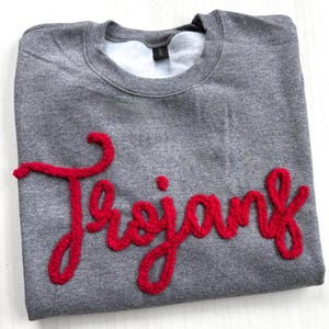 May include: A gray sweatshirt with the word "Trojans" in red, textured lettering across the front. The sweatshirt has a crew neck and a soft, white interior. The fabric appears to be a blend of cotton and polyester.