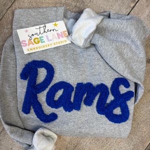 May include: A gray sweatshirt with the word "Rams" in bold, blue, textured letters. The sweatshirt has a white interior lining in the hood and a tag that reads "southern SAGE LANE EMBROIDERY STUDIO".