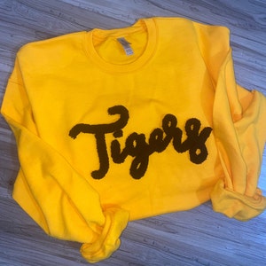 May include: A bright yellow sweatshirt with the word "Tigers" in a dark brown, textured font across the front. The sleeves are rolled up, and the sweatshirt is made of a soft material.