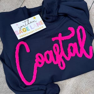 May include: Navy blue sweatshirt with the word "Coastal" in bright pink, fluffy lettering. A small tag with the text "southern SAGE LANE EMBROIDERY STUDIO" is attached. The sweatshirt is folded on a light wood surface.