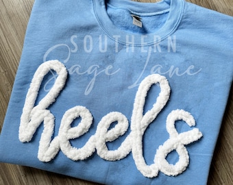 以下が含まれることがあります： 薄い青色のスウェットシャツに、白いふわふわの文字で「Heels」と書かれています。