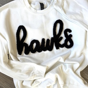 May include: A white sweatshirt with the word "hawks" in large, black, fuzzy letters across the chest. The long-sleeved top has a crew neck and a relaxed fit, suitable for casual wear.