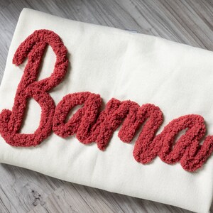 May include: A white throw pillow with the word "Bama" embroidered in red fluffy yarn.