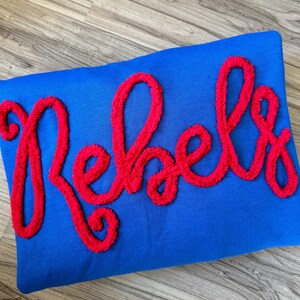May include: A blue pillow with the word "Rebel" spelled out in red fuzzy letters.