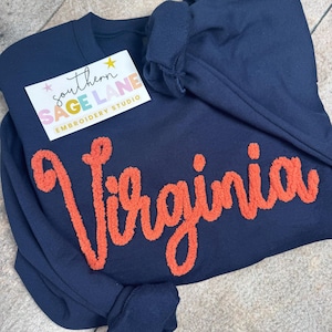 May include: Navy blue sweatshirt with the name "Virginia" embroidered in orange, cursive lettering. A small white card with the text "southern SAGE LANE embroidery studio" is on the upper left. The sweatshirt's sleeve is folded over.