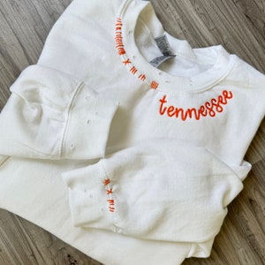 Może przedstawiać: Biała bluza z napisem "tennessee" wyhaftowanym pomarańczową czcionką. Dekolt i mankiety zdobią ozdobne pomarańczowe przeszycia. Bluza jest złożona na drewnianej powierzchni.