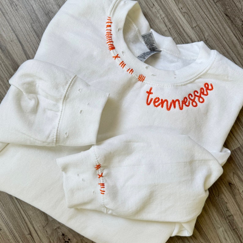 Może przedstawiać: Biała bluza z napisem "tennessee" wyhaftowanym pomarańczową czcionką. Dekolt i mankiety zdobią ozdobne pomarańczowe przeszycia. Bluza jest złożona na drewnianej powierzchni.