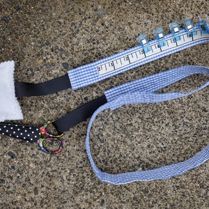 May include: A sewing kit with a white pincushion, blue pins, black and white polka dot scissors, and a blue and white gingham measuring tape with blue clips. The tape has inch markings.