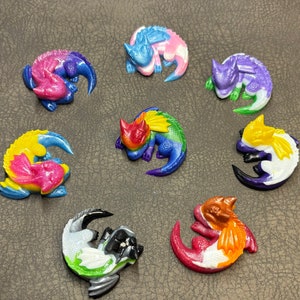 Puede incluir: Ocho pequeñas figuritas de dragón impresas en 3D y de colores. Cada dragón está enroscado en una posición de sueño. Los dragones están pintados en varios colores, incluyendo azul, morado, rosa, amarillo, verde, naranja, blanco y negro.