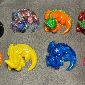 May include: Eight colourful, glittery dinosaur figurines in various colours, including black, orange, yellow, blue, and red. Each figurine is a unique design, with a textured surface and a glossy finish.