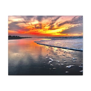 Puede incluir: Una vibrante puesta de sol sobre una playa con un muelle en la distancia. El cielo es una mezcla de naranja, rosa y morado, y el agua refleja los colores. La arena está mojada y hay pequeñas olas que llegan a la orilla.