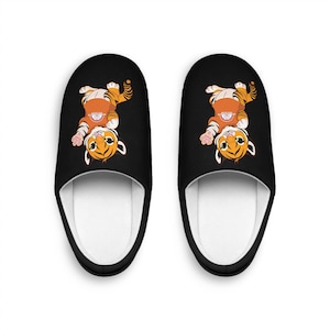 Könnte beinhalten: Paar schwarze Hausschuhe mit einem Cartoon-Tiger-Design. Der Tiger ist orange und weiß, mit einem lächelnden Gesicht und gestreiftem Schwanz. Die Hausschuhe haben eine weiße Innenseite und eine schwarze Außenseite.