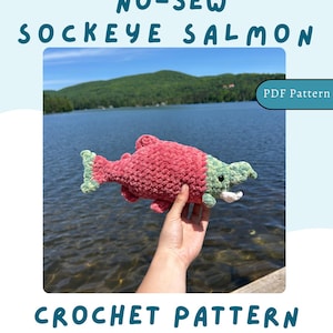May include: A handmade, no-sew crochet Sockeye Salmon in shades of red and green. The fish is held up against a backdrop of a lake and mountains. The image includes the text "CROCHET PATTERN" and "NO-SEW SOCKEYE SALMON".
