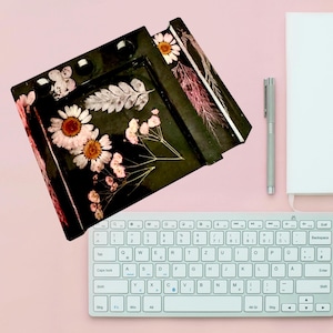 Puede incluir: Organizador de escritorio de resina negra con flores secas incrustadas, incluyendo margaritas y delicadas flores rosas. El organizador tiene múltiples compartimentos y está sobre una superficie rosa con un teclado, bolígrafo y cuaderno.