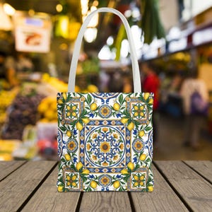 May include: A white tote bag featuring a repeating pattern of yellow lemons, green leaves, and blue and yellow floral tile motifs. The bag has two white fabric handles and a square-shaped body with a detailed, symmetrical Mediterranean-inspired design.