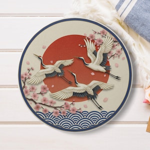 Puede incluir: Plato decorativo redondo con un diseño de inspiración japonesa. Presenta tres grullas blancas en vuelo frente a un sol rojo, con flores de cerezo rosas y un borde con patrón de olas azules. El plato tiene un diámetro de aproximadamente 25 cm.