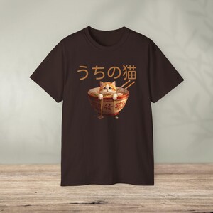 May include: Brown t-shirt featuring a graphic of a cat in a ramen bowl with chopsticks. The text "うちの猫" is above the bowl. The cat is orange and white, and the bowl is red and gold.