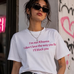 Puede incluir: Camiseta blanca con texto rosa: "I'm not Rihanna, I don't love the way you lie, I'll block you". La persona lleva gafas de sol negras, un collar plateado y un cinturón negro. Hay graffiti en el fondo.