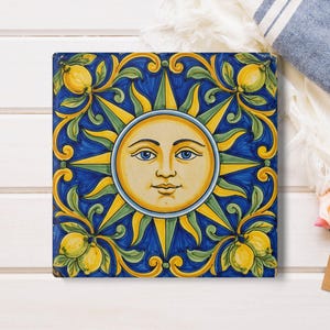 Puede incluir: Azulejo de cerámica cuadrado con un diseño de sol. El sol tiene una cara sonriente con ojos azules y está rodeado de rayos amarillos. El fondo es azul oscuro con detalles florales y de limón amarillos y verdes.