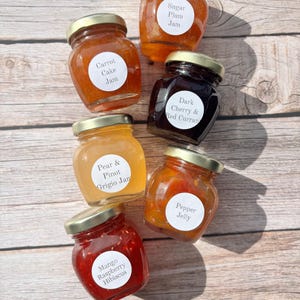 May include: A collection of six small glass jars filled with colorful jams and jellies. Each jar has a gold lid and a white label with the flavor name: Sugar Plum, Carrot Cake, Dark Cherry & Red Currant, Pepper Jelly, Mango Raspberry Hibiscus, and Pear & Pinot Grigio.