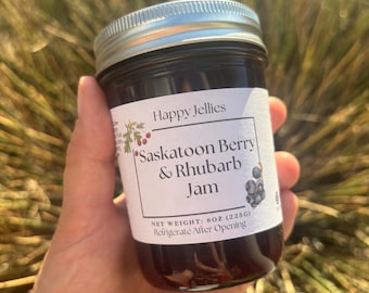 May include: A jar of Saskatoon Berry & Rhubarb Jam by Happy Jellies. The label indicates a net weight of 8 oz (225g). The jar is filled with dark jam and has a silver lid. The label also says to refrigerate after opening.