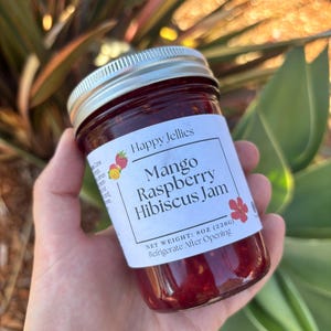 May include: A jar of Mango Raspberry Hibiscus Jam by Happy Jellies. The label includes the text "NET WEIGHT: 8OZ (226g) Refrigerate After Opening". The jam is a deep red color, and the jar has a silver lid.