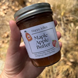 May include: A glass jar of Happy Jellies Maple Apple Butter with a silver metal lid. The white label features a small illustration of a maple syrup bottle and a red apple, with the text Maple Apple Butter, Net Weight: 8oz (226g), and Refrigerate After Opening.