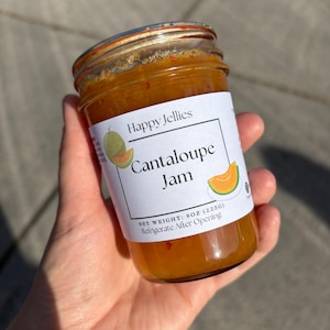 May include: A jar of cantaloupe jam from Happy Jellies. The label features a cantaloupe illustration. The jar contains a bright orange jam and is held in a hand.