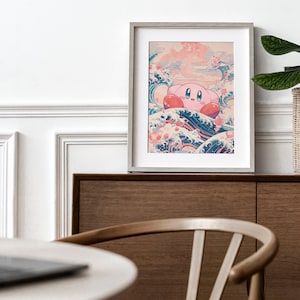 May include: A framed print of a pink cartoon character, Kirby, riding a large wave with a blue and white design. The wave is surrounded by pink flowers and a pink sky. The print is framed in a light gray frame and is sitting on a wooden shelf.