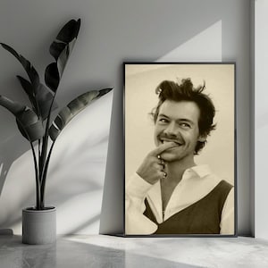 Può includere: Ritratto in bianco e nero incorniciato di una persona sorridente con i capelli scuri, che indossa una camicia bianca e un gilet scuro. La persona si tocca il viso con un dito. Una pianta in vaso è a sinistra.