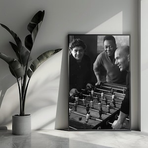 Könnte beinhalten: Schwarz-Weiß-Druck mit drei Männern beim Tischfußball. Das Bild ist vor einer neutralen Wand platziert, mit einer Topfpflanze im Vordergrund. Ein klassisches Dekorationselement.